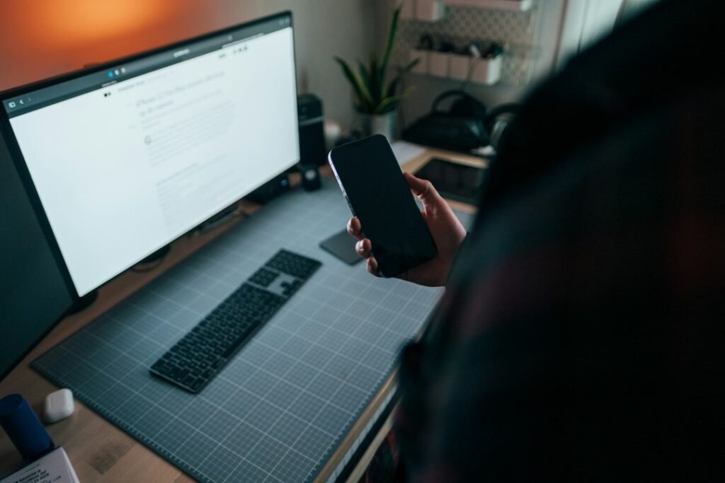 Person holding a smartphone beside a laptop at a desk