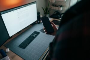 Person holding a smartphone beside a laptop at a desk